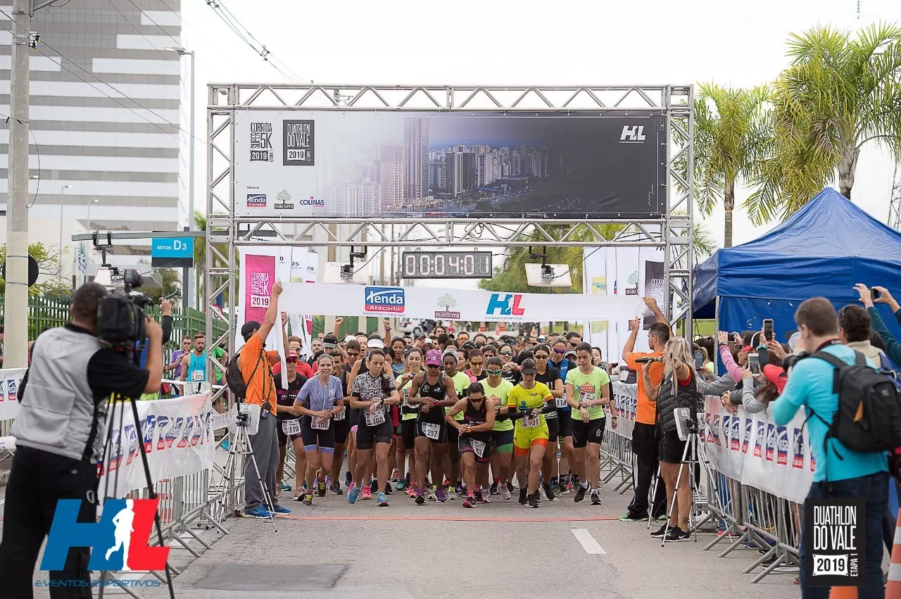 Foto mostra corredores da corrida de rua Duathlon, em SJC, em 2019, com uma placa em cima, cinegrafistas em volta e pessoas tirando foto.