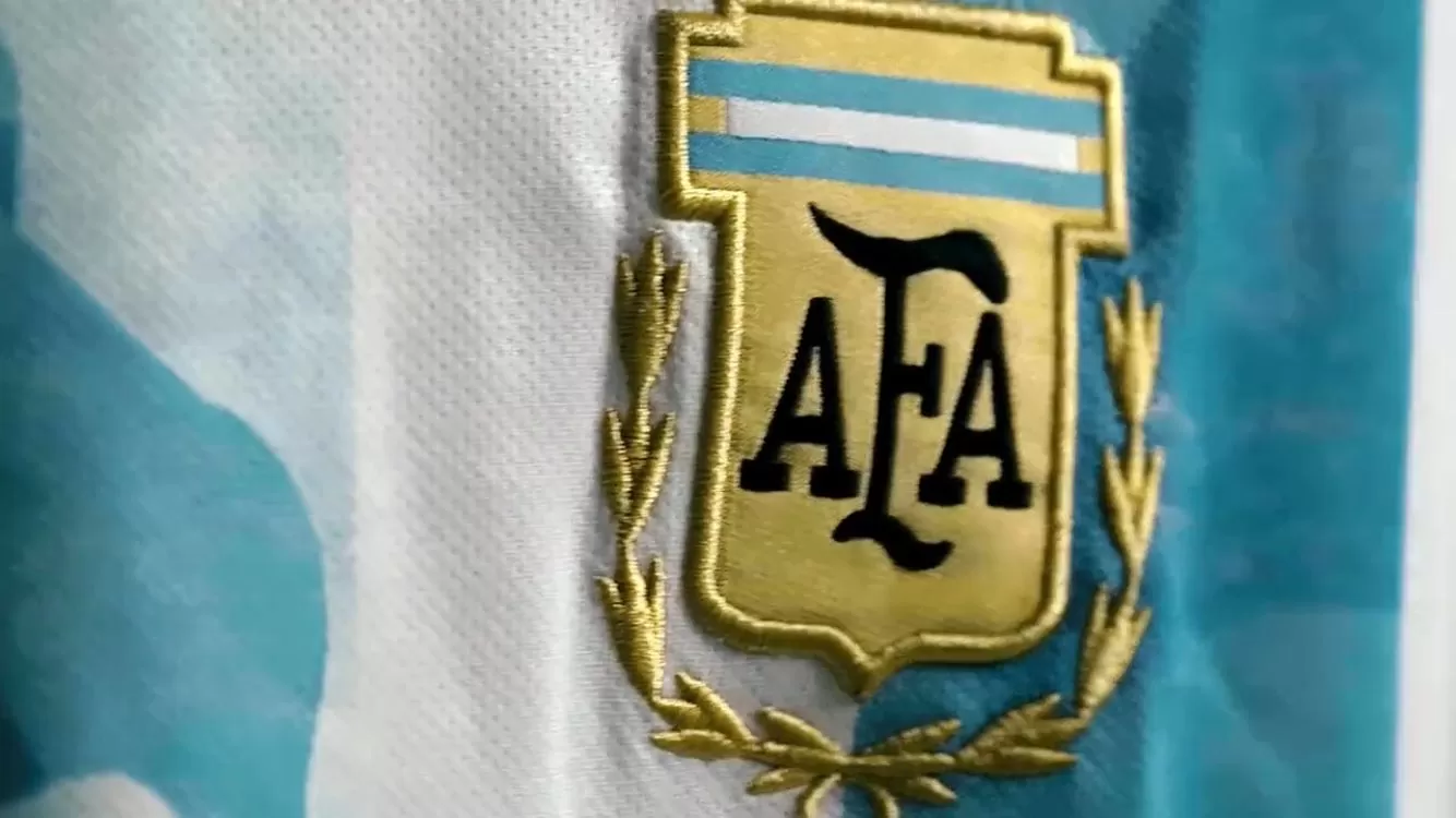 Close em camisa da seleção argentina listrada de azul e branco e exibindo o escudo da AFA ao lado esquerdo do peito.