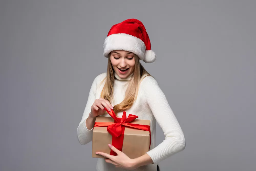 Mulher branca, loira, com uma blusa branca, segurando uma caixa marrom, com um laço vermelho, vestindo uma touca de Papai Noel; foto ilustra a matéria sobre dicas de presentes de Natal em SJC (São José dos Campos), segundo a Humanitarian Calçados.