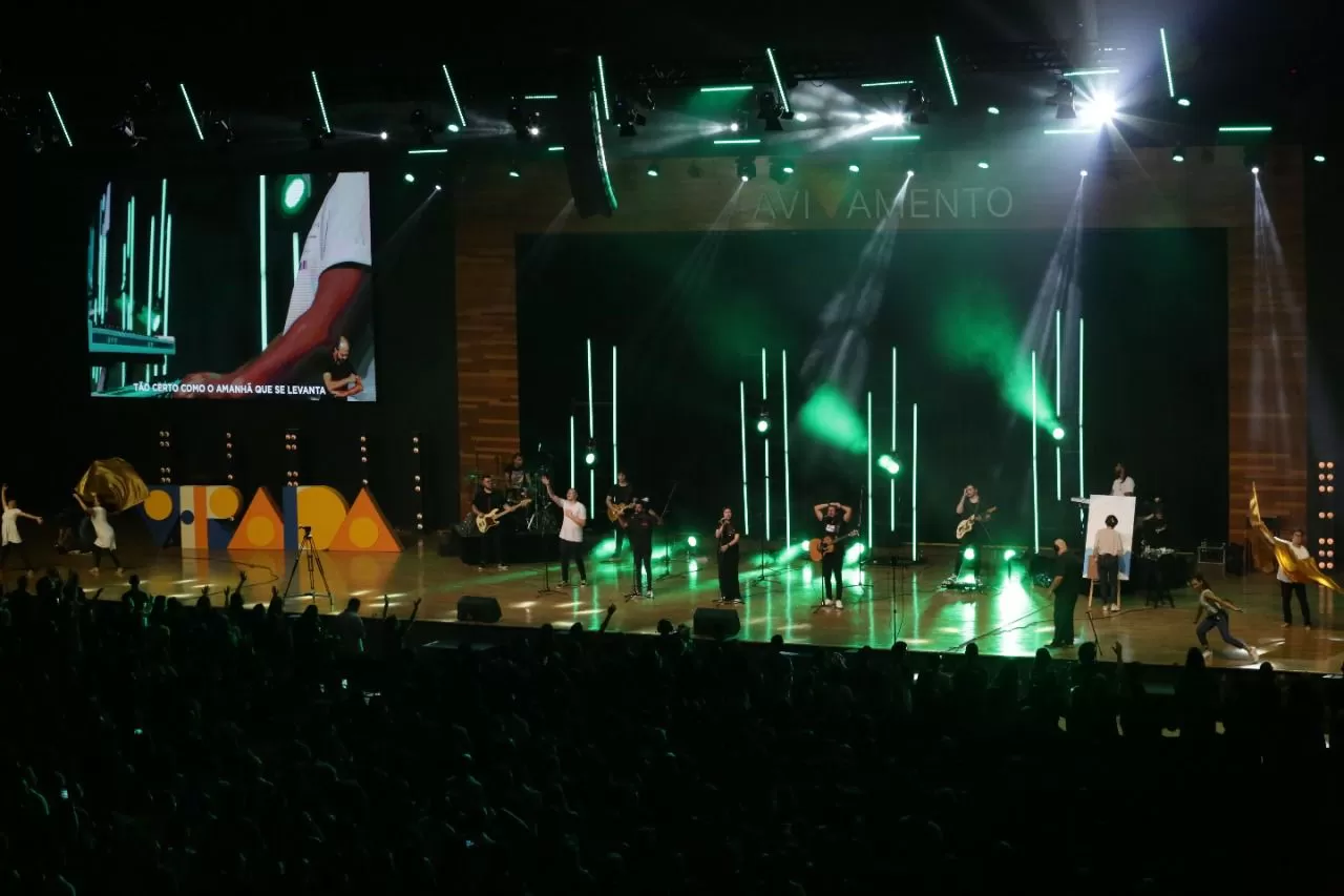 Palco da Semana da Virada, da Igreja da Cidade de SJC, com músicos tocando, plateia lotada, telão ao fundo, luzes verdes e ambiente escuro.