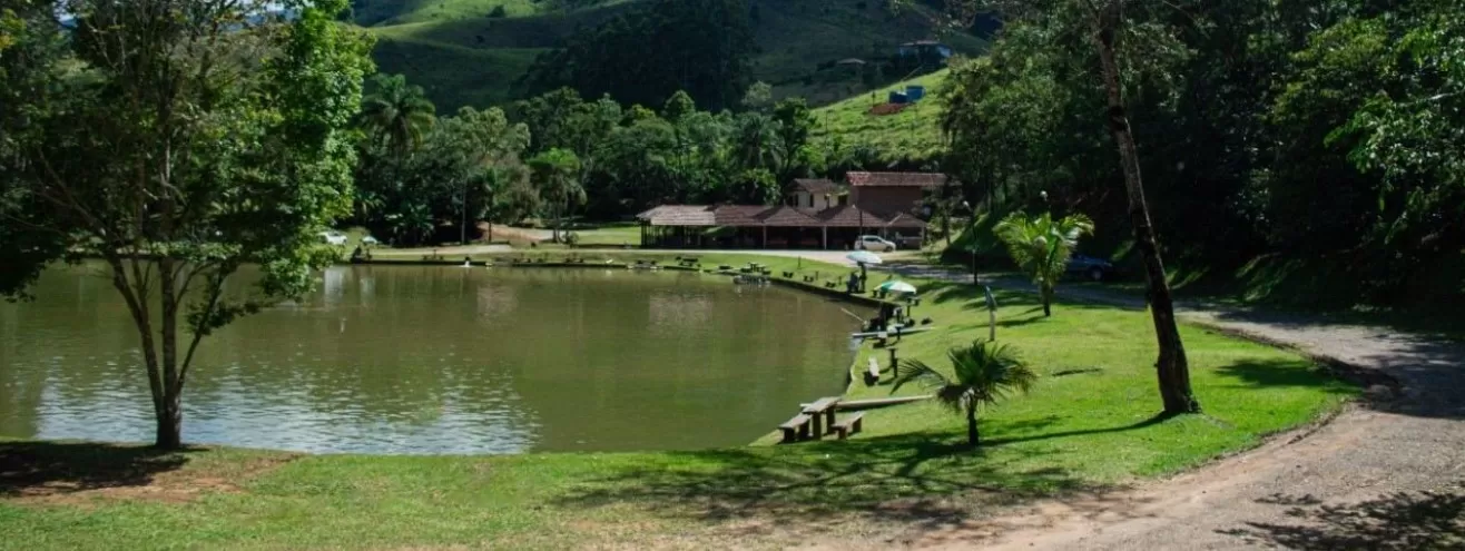 Parte verde do Pesqueiro Pantanal Pesca e Lazer, em São José dos Campos, com um lado no meio, banquinhos ao redor, muitas árvores e uma construção ao fundo, em madeira.