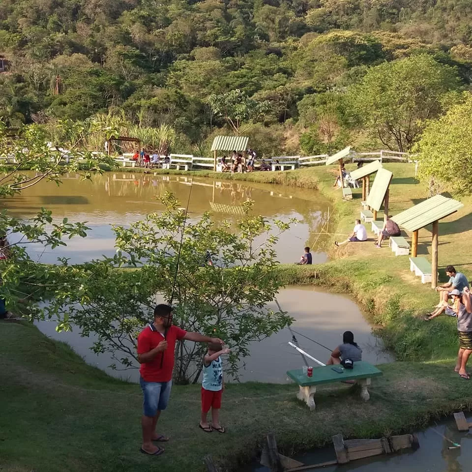 Foto mostra área verde, lado ao fundo e pessoas em volta pescando no Pesqueiro Recanto do Sol, em São José dos Campos.