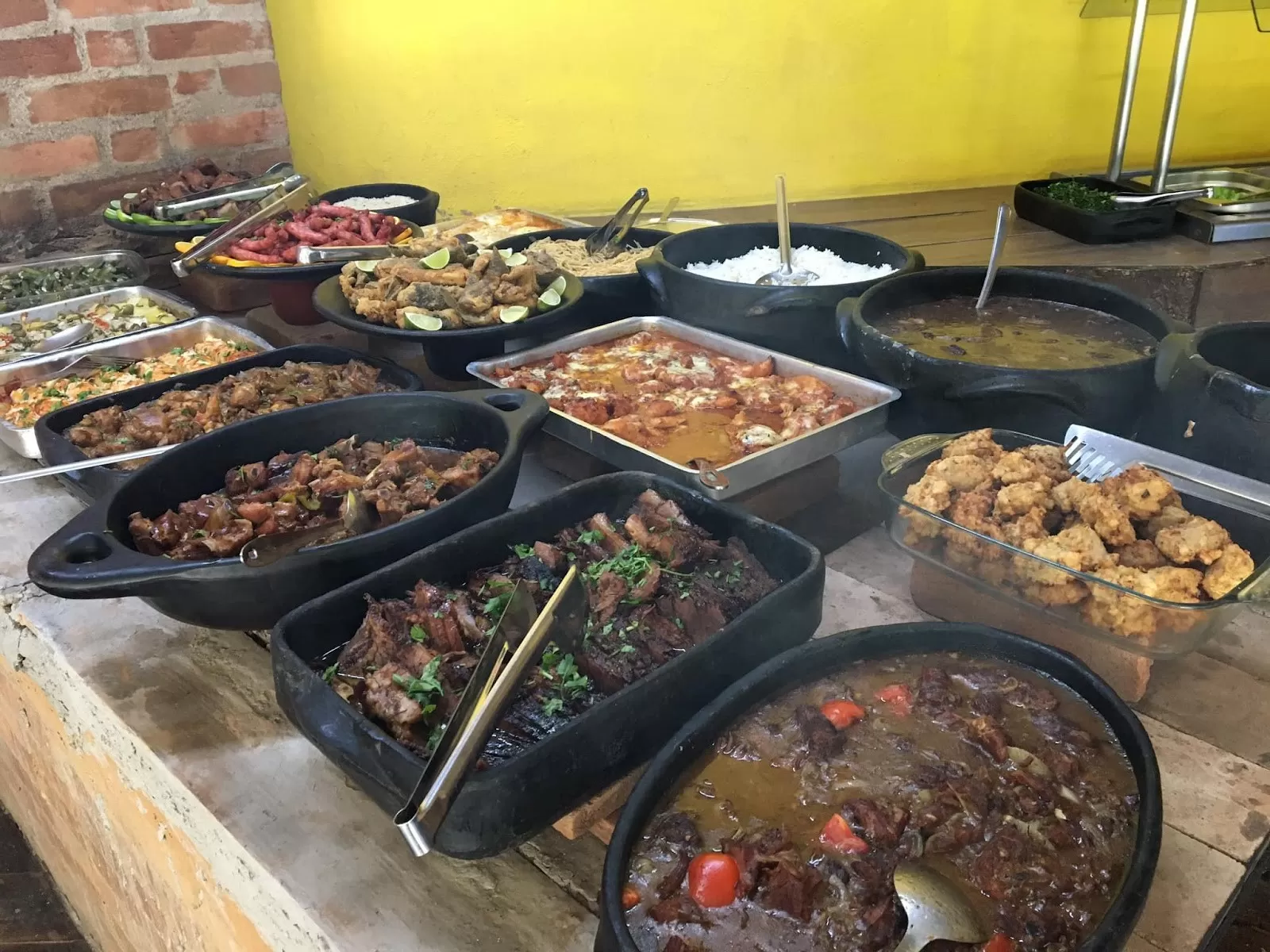 Diversas comidas em cima de uma mesa de madeira, panela de barro preto e uma parede amarela ao fundo.