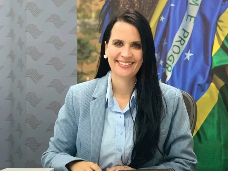 Retrato de Letícia Aguiar com terno e camisa azul, em frente a uma bandeira do Brasil, com cabelo preto e sorrindo; foto ilustra matéria sobre presidência do movimento Mulheres Progressistas.