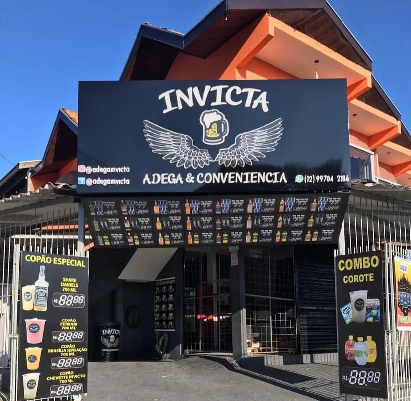 Adega Invicta é outra opção de adegas da zona sul de SJC; foto mostra fachada com placa preta, letras brancas, com portas abertas e desenhos de bebidas na frente. Lista completa inclui opções abertas 24 horas.