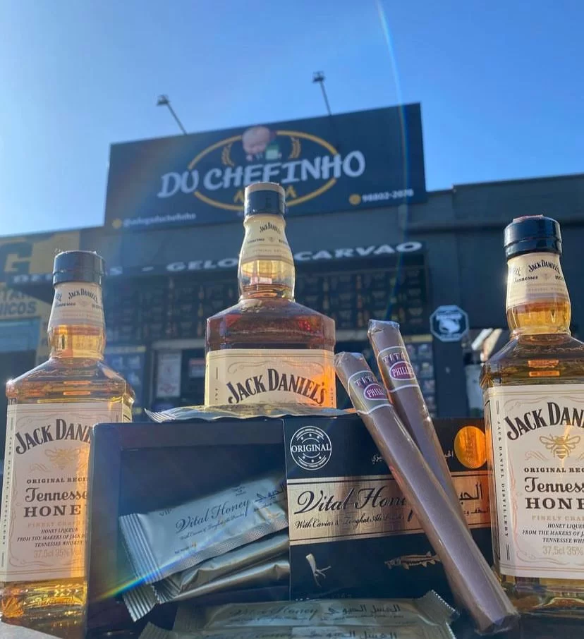 Mais uma opção de adega na zona sul de SJC, a Adega Du Chefinho; foto mostra placa preta com letras brancas e amarelas, com uma vista de baixo para cima, um céu azul ao fundo e bebidas na parte da frente, incluindo Jack Daniel's e tabacos.