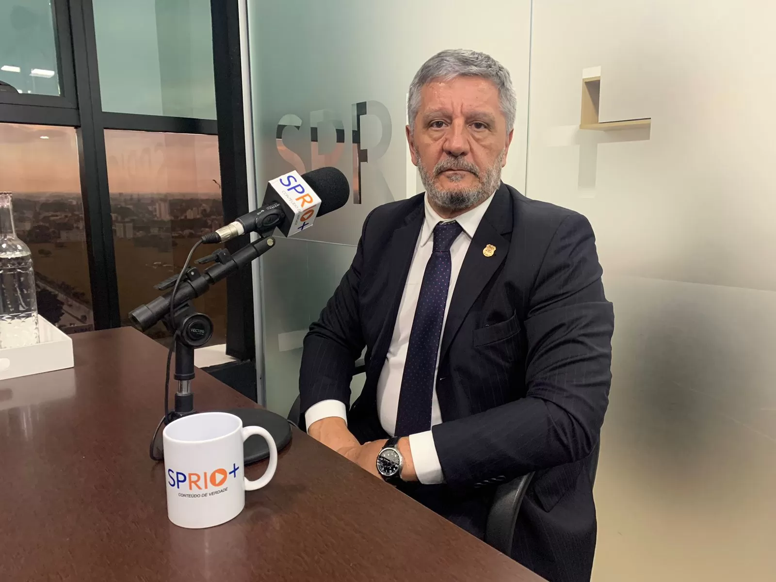 Delegado Waldir Covino, da Polícia Civil do Vale do Paraíba, com terno preto, gravata preta e camisa branca, sentado em uma cadeira, em frente a um microfone e uma mesa de madeira, com um vidro ao fundo.