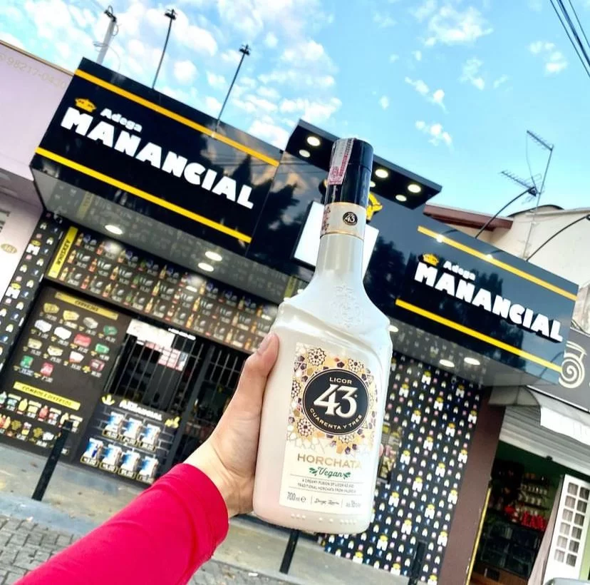 Adega Manancial é uma das melhores adegas da Zona Sul de SJC; foto mostra uma mão com roupa rosa segurando uma garrafa de bebida em frente a fachada da empresa, com um céu azul ao fundo.