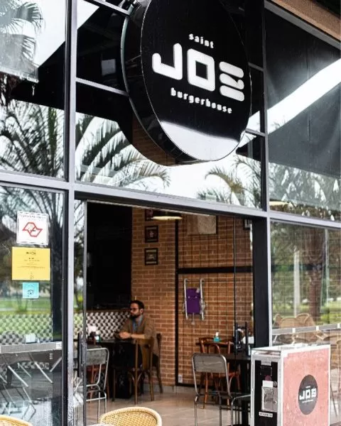 Saint Joe Burgerhouse, com uma placa preta em cima, com letras brancas, detalhes em vidro e uma pessoa sentada em uma cadeira de madeira ao fundo, enquanto come algo; hamburgueria está no Jardim Aquarius, em São José dos Campos.