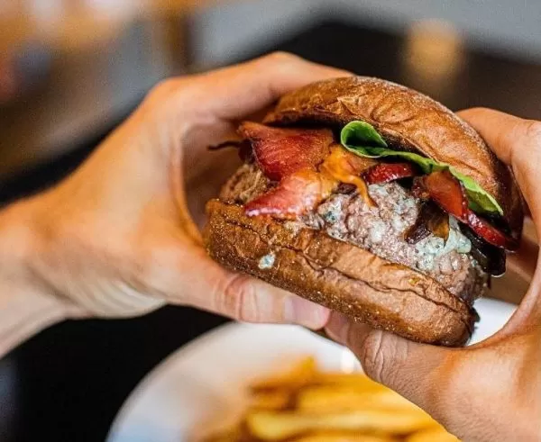 Lanche do Saint Joe Burgerhouse, com alface, tomate e carne, enquanto uma pessoa o segura, com um prato branco em baixo, com batata frita e o fundo desfocado.