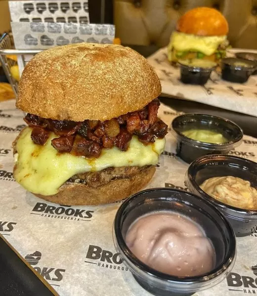 Lanche do Brooks, na Avenida Anchieta, em São José dos Campos, com um pão, bacon, queijo e carne, em cima de uma mesa, com molhos ao lado; foto ilustra matéria sobre hamburguer artesanal em SJC.