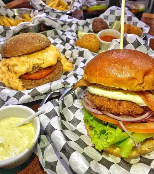 Lanches do Burguin, hamburgueria artesanal em São José dos Campos (SJC), em cima de um papel preto e branco, com molhos em volta e potes de batata frita ao fundo.