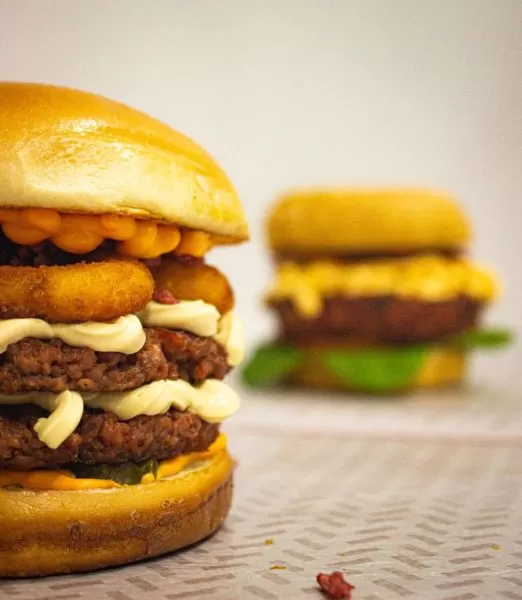 Uma opção de lanche no Caravegana, em São José dos Campos; foto mostra pão, hamburguer, molho e cheddar, em cima de uma mesa com toalha branca; ao fundo há outro lanche, mas desfocado.