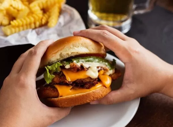 Mão segurando lanche do Container Burger, em SJC, em cima de um prato branco e está apoiado em uma mesa preta, com batata frita ao fundo e um copo de cerveja.