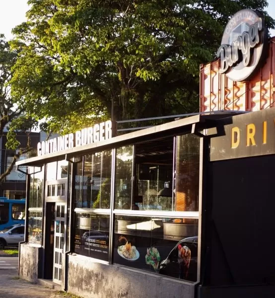 Fachada do Container Burger, no Jardim Esplanada, em São José dos Campos, com uma árvore na parte de cima da foto, uma placa com o nome da empresa e detalhes em vidro.
