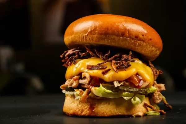 Foto ilustra matéria sobre hamburguer artesanal em SJC (São José dos Campos), com um fundo preto, o alimento na frente, bem recheado, com alface e queijo cheddar.
