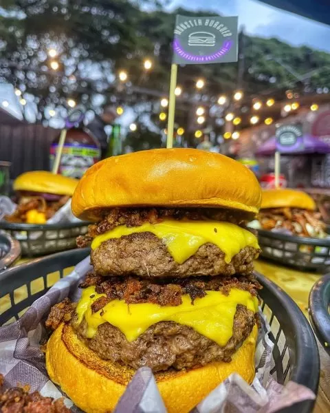 Lanche do Monster Burger, no Parque Indsustrial, com muito queijo e carne; ao fundo há o cenário do local, com luzes acesar e árvores; foto ilustra matéria sobre hamburguer artesanal em SJC.