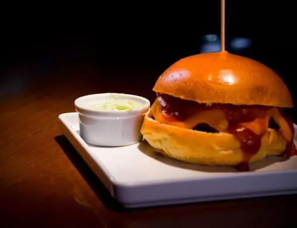 Opção no Paulista Burger & Bar, com um lanche em cima de um prato branco, com um palito espetato em cima, ao lado de um pote de molho, com um fundo preto.