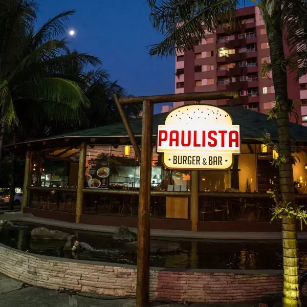 Fachada do Paulista Burger & Bar, localizado no Jardim Aquarius, em São José dos Campos, com detalhes em madeira, uma placa iluminada, árvores ao redor e um lago no centro.