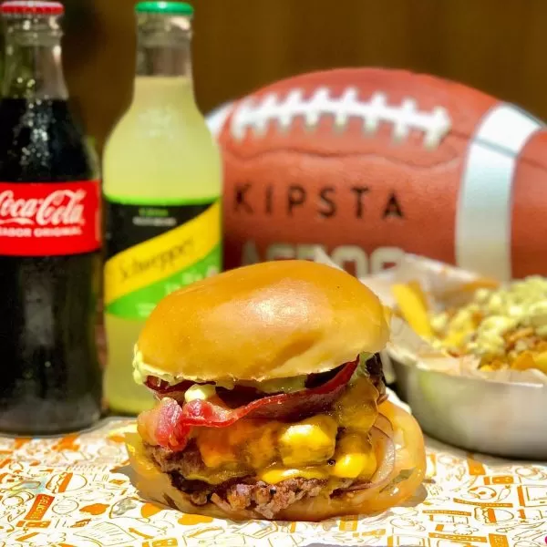 Lanche do PlayOff Burger, uma das opções de hamburger artesanal em SJC (São José dos Campos), no bairro Aquarius; foto mostra um lanche em frente a bebidas, uma porção de batata e uma bola de futebol americano ao fundo.