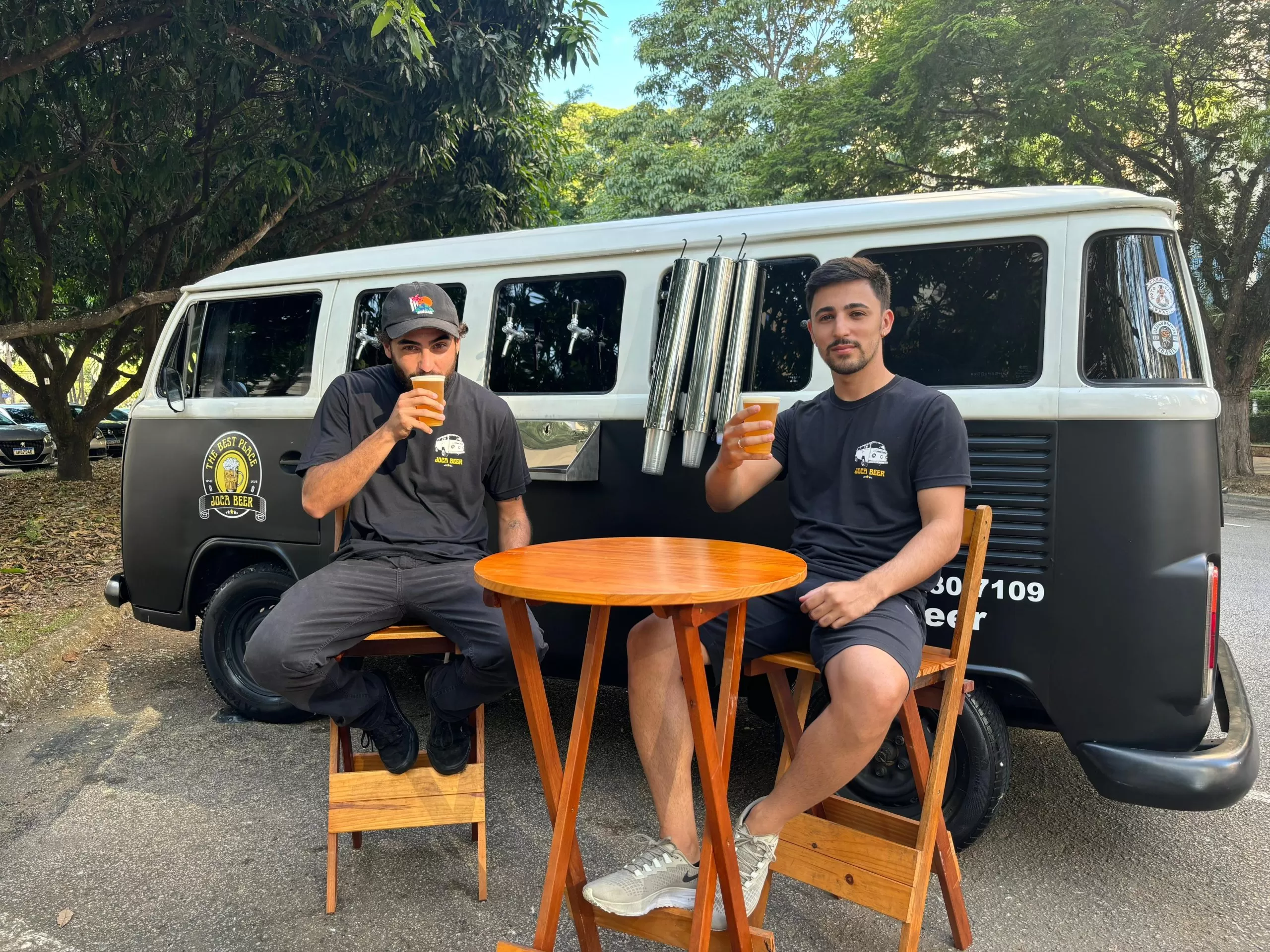 matheus bebendo cerveja sentado ao lado de gabriel, que segura copo de cervej; os dois estão entados em cadeiras e ao lado de mesinha montada em frente á kombi da joca beer