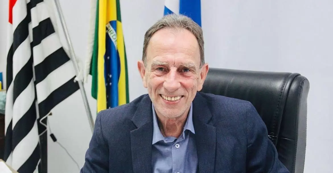 piriquito, prefeito de aparecida, em foto sorrindo sentado em sala com bandeira do brasil aos fundos