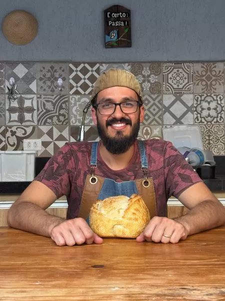 junior sorrisndo, com boina, sentado na mesa e na frente um pão italiano